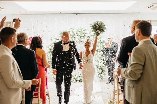 Newlywed couple walk down the aisle through falling confetti as wedding guests applaud.