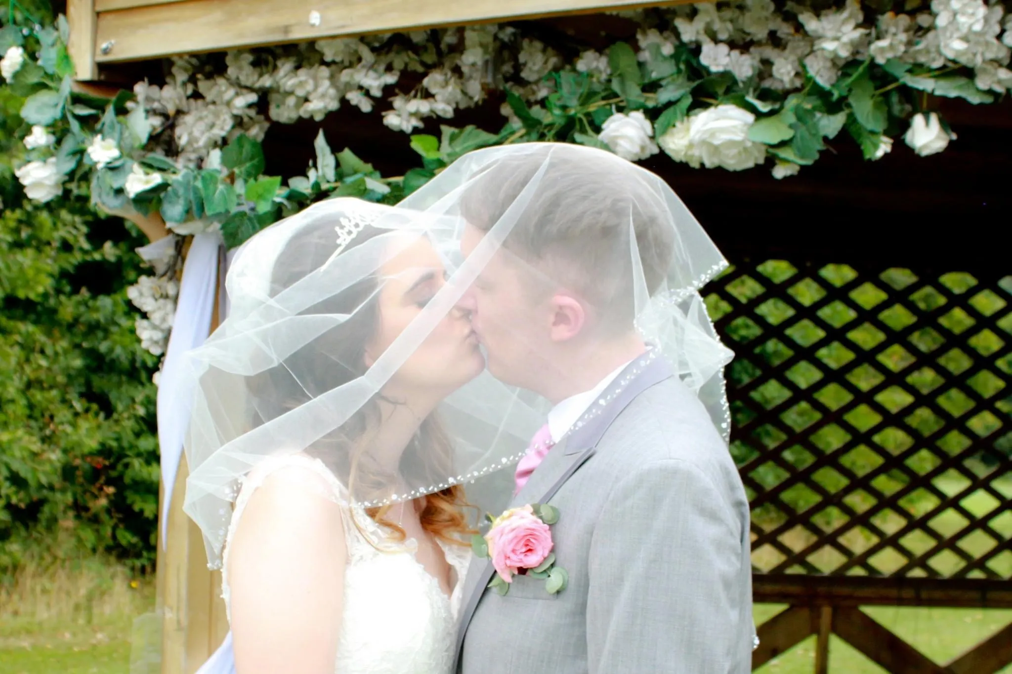 Bride and groom kissing beneath a floral wedding arch, covered by the bride's veil.