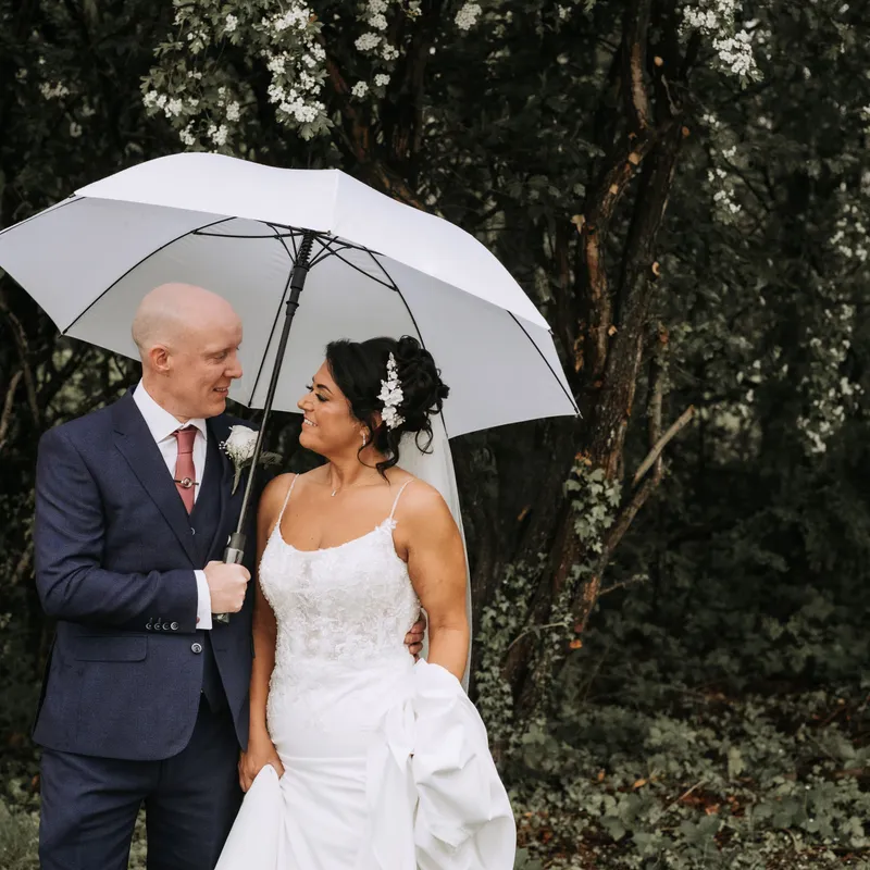 Bride and groom smiling beneath a white umbrella in a leafy woodland setting.