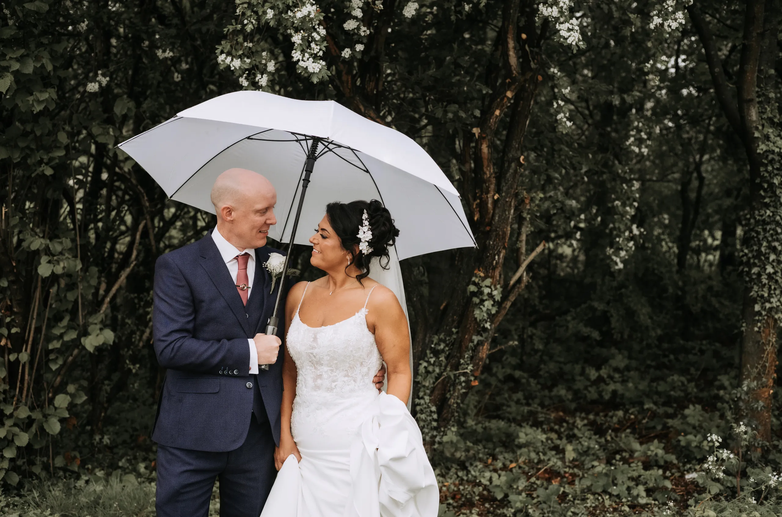 Bride and groom smiling beneath a white umbrella in a leafy woodland setting.