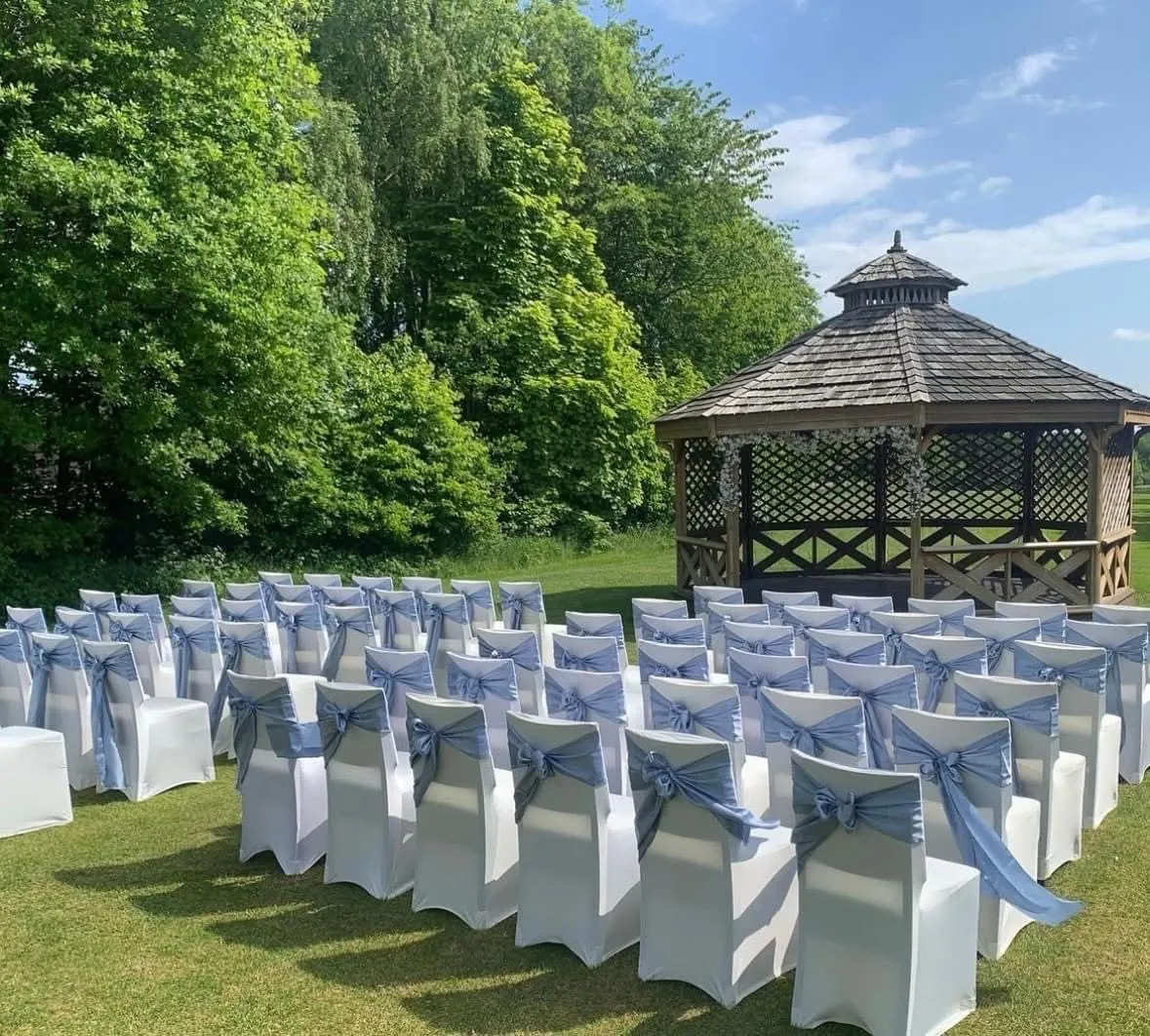 Outdoor wedding ceremony seating with white chair covers and pale blue bows beside a wooden gazebo