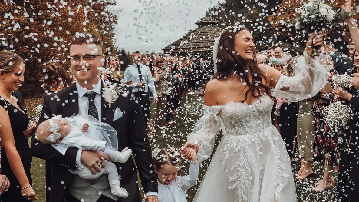 Joyful newlyweds walk through a shower of white confetti with their young children and wedding guests outdoors.