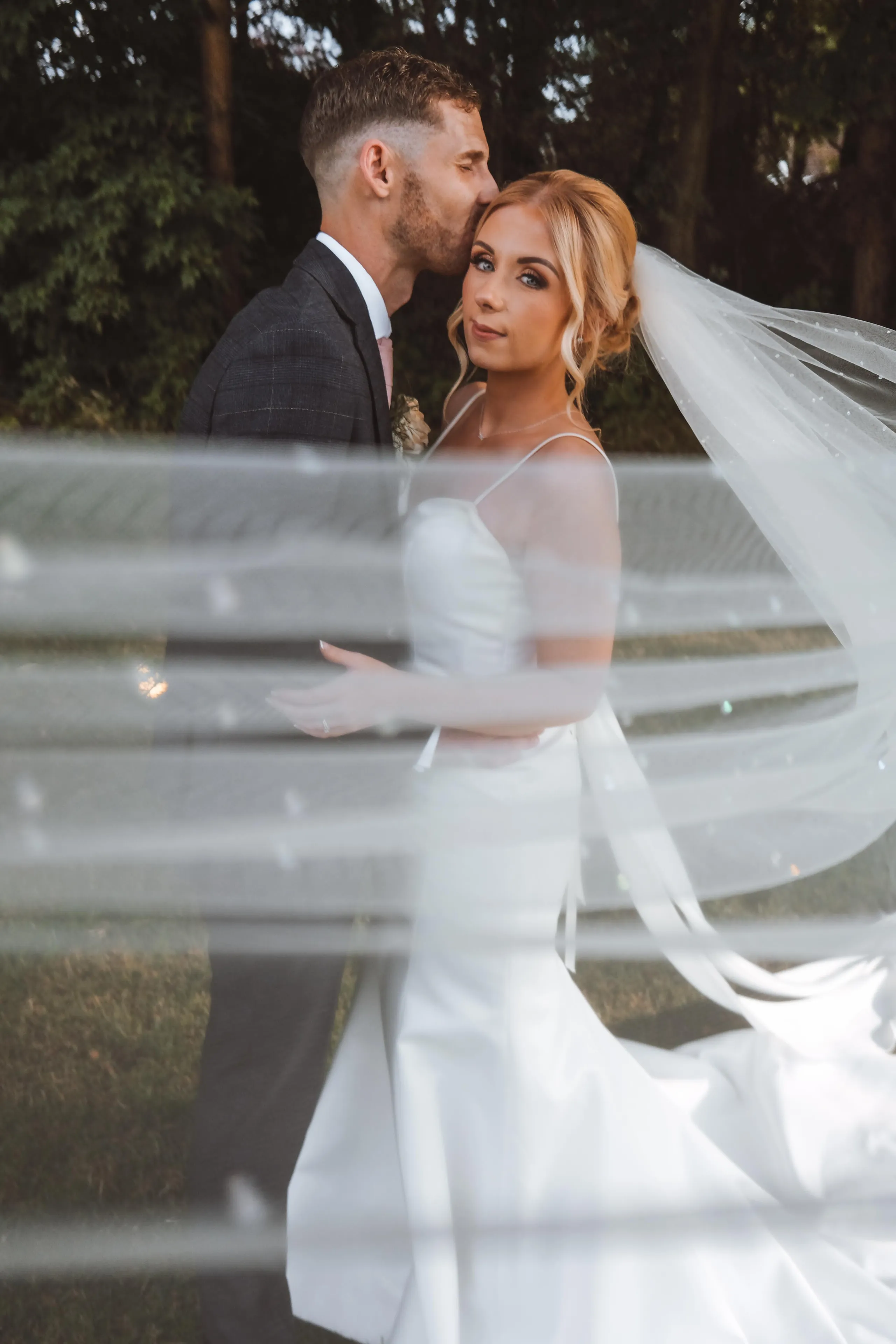 Groom kissing bride's forehead as they embrace outdoors, photographed through a translucent veil.