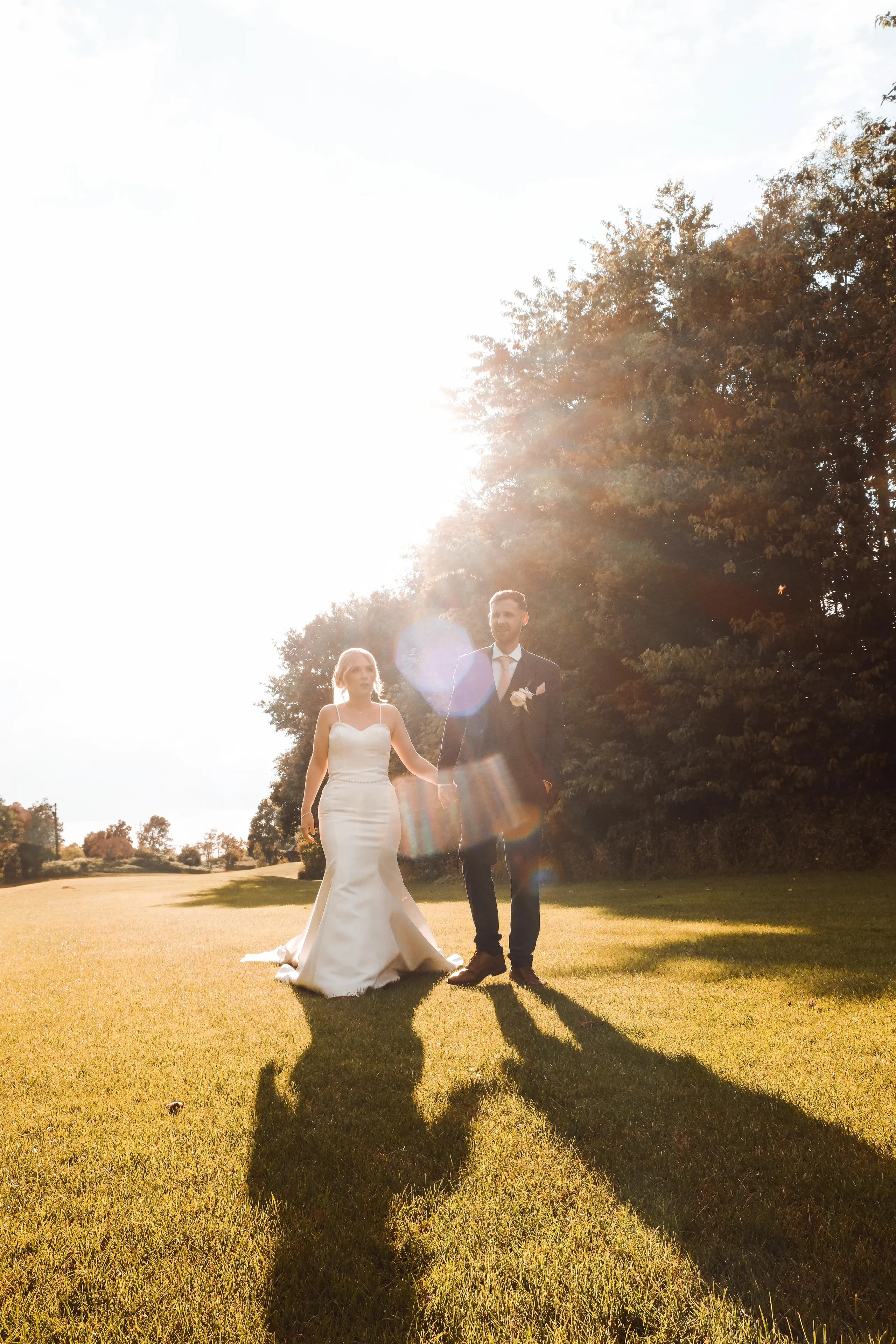 Newlywed couple holding hands and walking across a sunlit lawn at golden hour