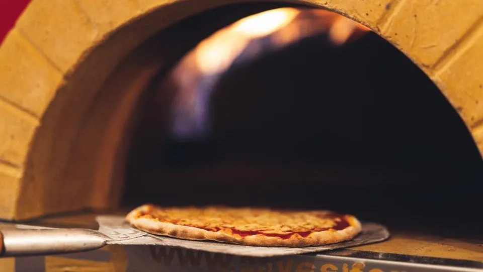 Pizza on a metal peel being placed into a glowing wood-fired brick oven