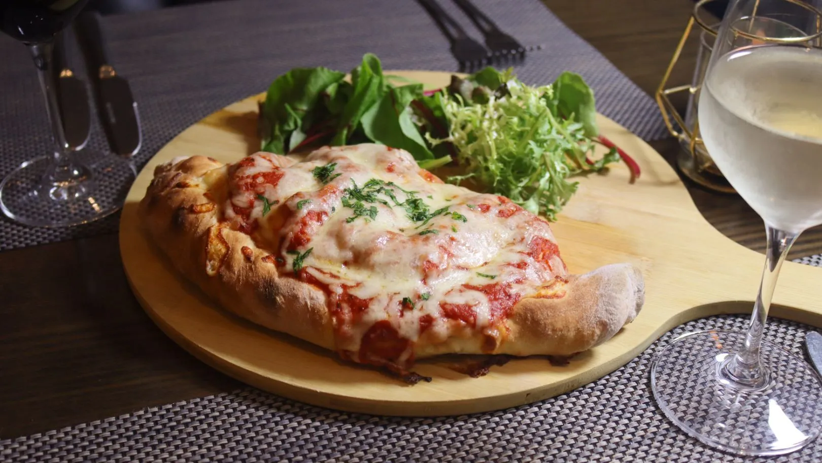 Cheese-topped baked pizza served on a wooden board with mixed salad and wine glasses
