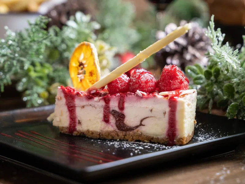 Slice of raspberry cheesecake topped with fresh raspberries, berry sauce and white chocolate on a black plate