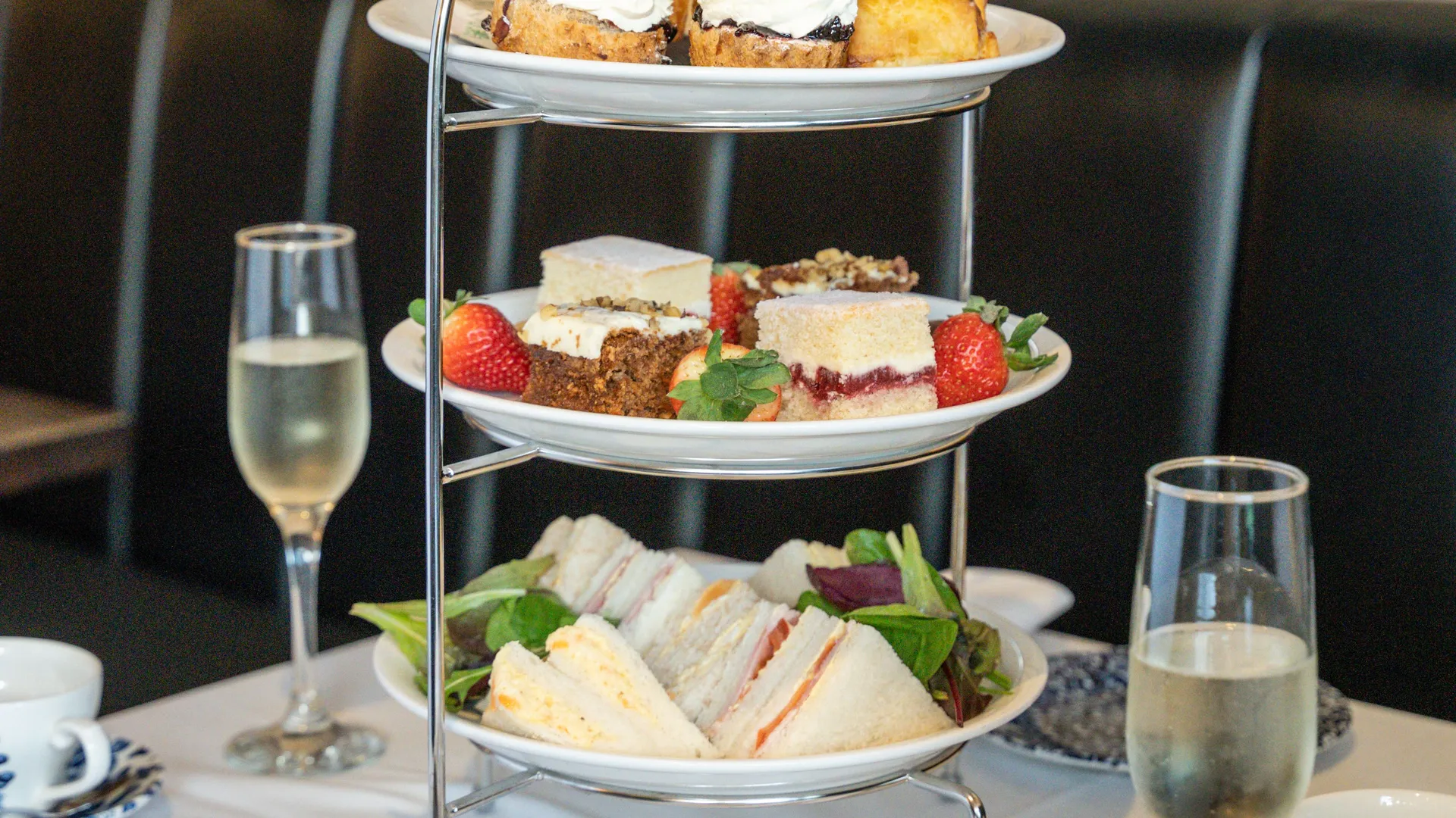 Three-tier afternoon tea stand with sandwiches, cakes and scones beside champagne glasses on a restaurant table.