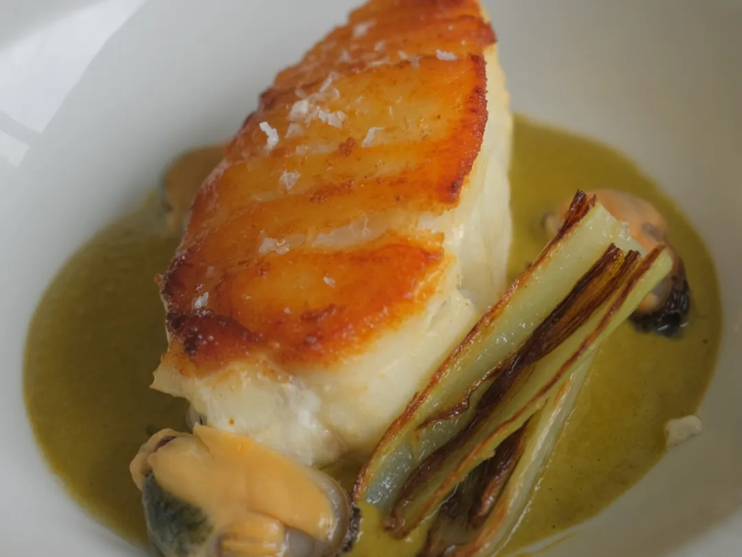 Seared fish fillet with mussels, grilled leek, and green sauce in a white bowl
