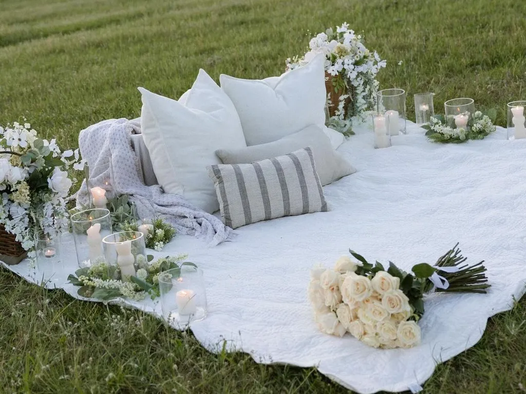 Romantic picnic setup on a blanket with pillows, candles, flowers, and a bouquet of white roses in a grassy field