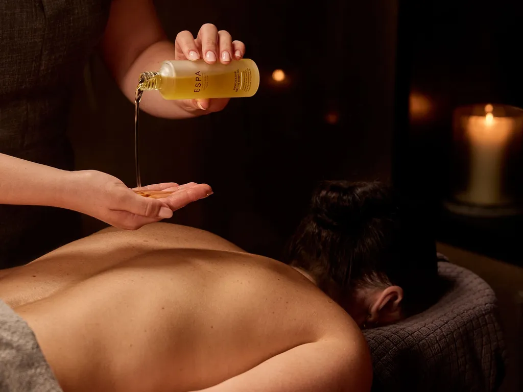 Massage therapist pouring oil onto their hand during a spa treatment with a candle burning in the background.