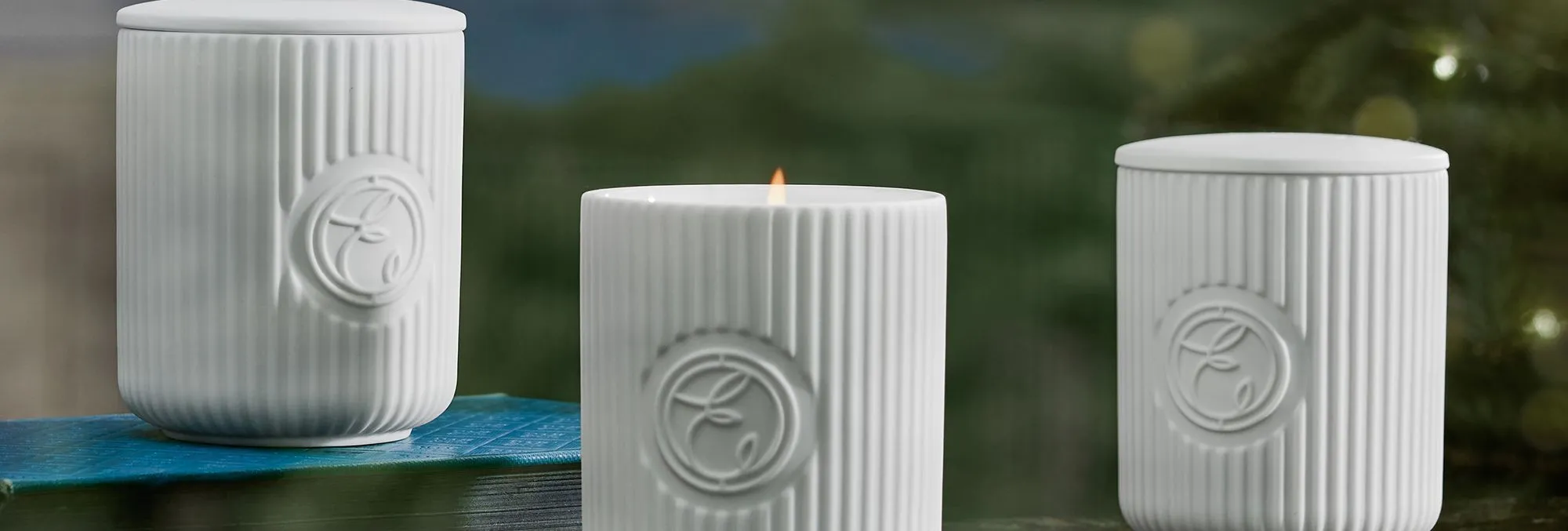 Three white ribbed ceramic candles, one lit, on a marble surface with a scenic background.