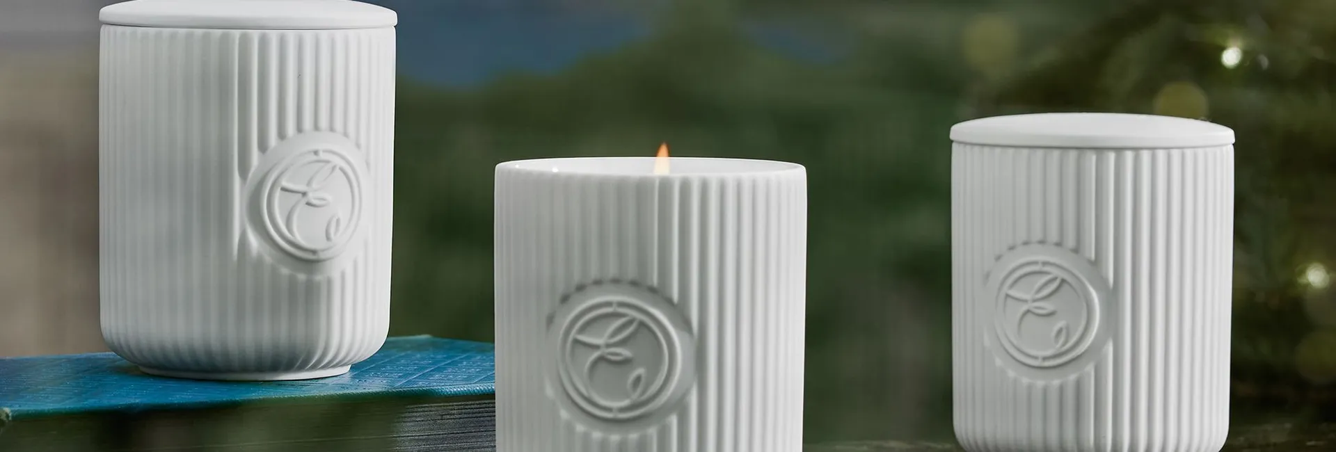 Three white ribbed ceramic candles, one lit, on a marble surface with a scenic background.