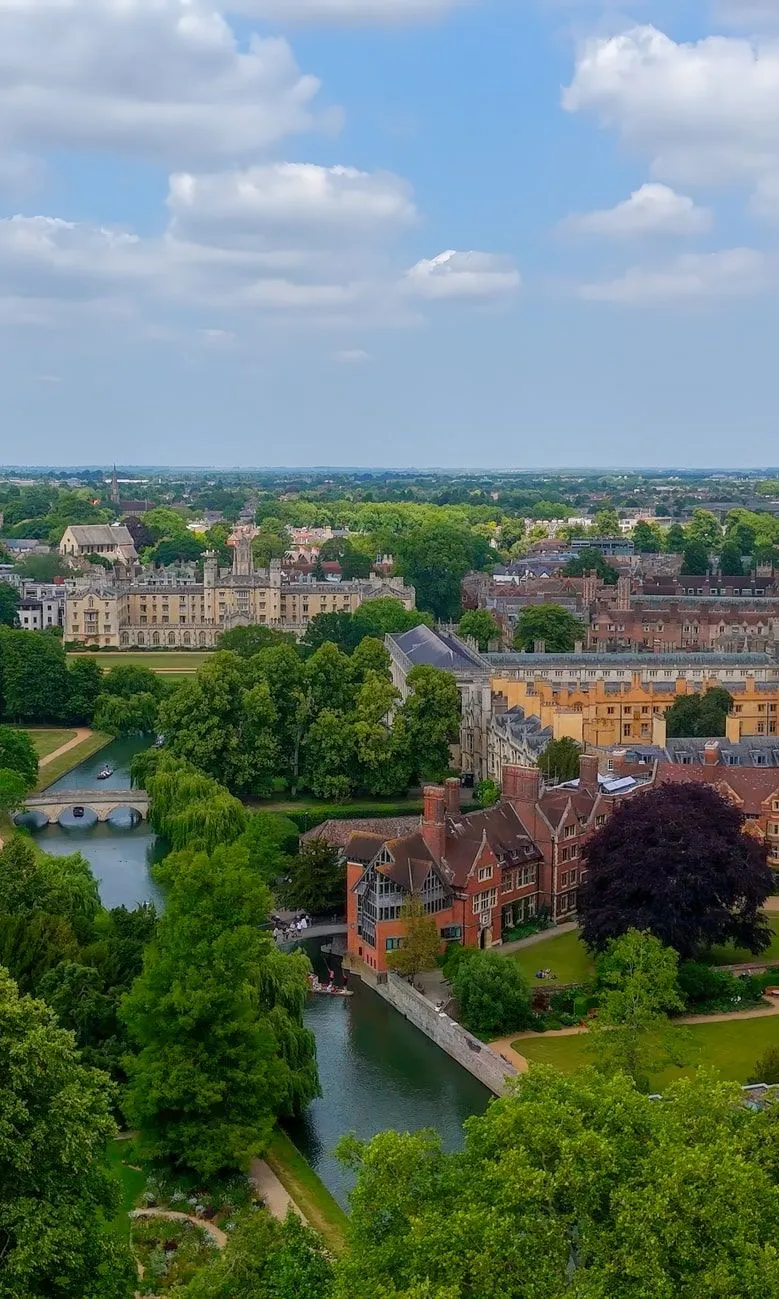 Aerial view of Cambridge University with historic college buildings, the River Cam, and lush green trees under a partly cloudy sky.