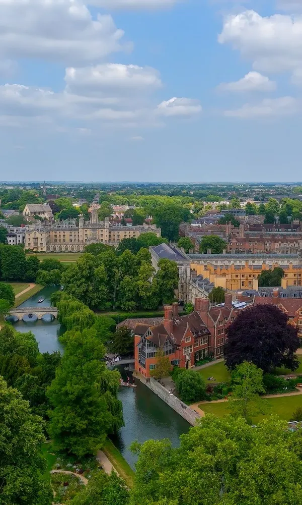 Aerial view of Cambridge University with historic college buildings, the River Cam, and lush green trees under a partly cloudy sky.