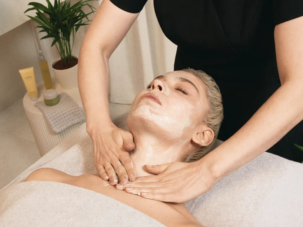 Person receiving a facial treatment at a spa, with a mask applied to the face and a therapist massaging their neck.