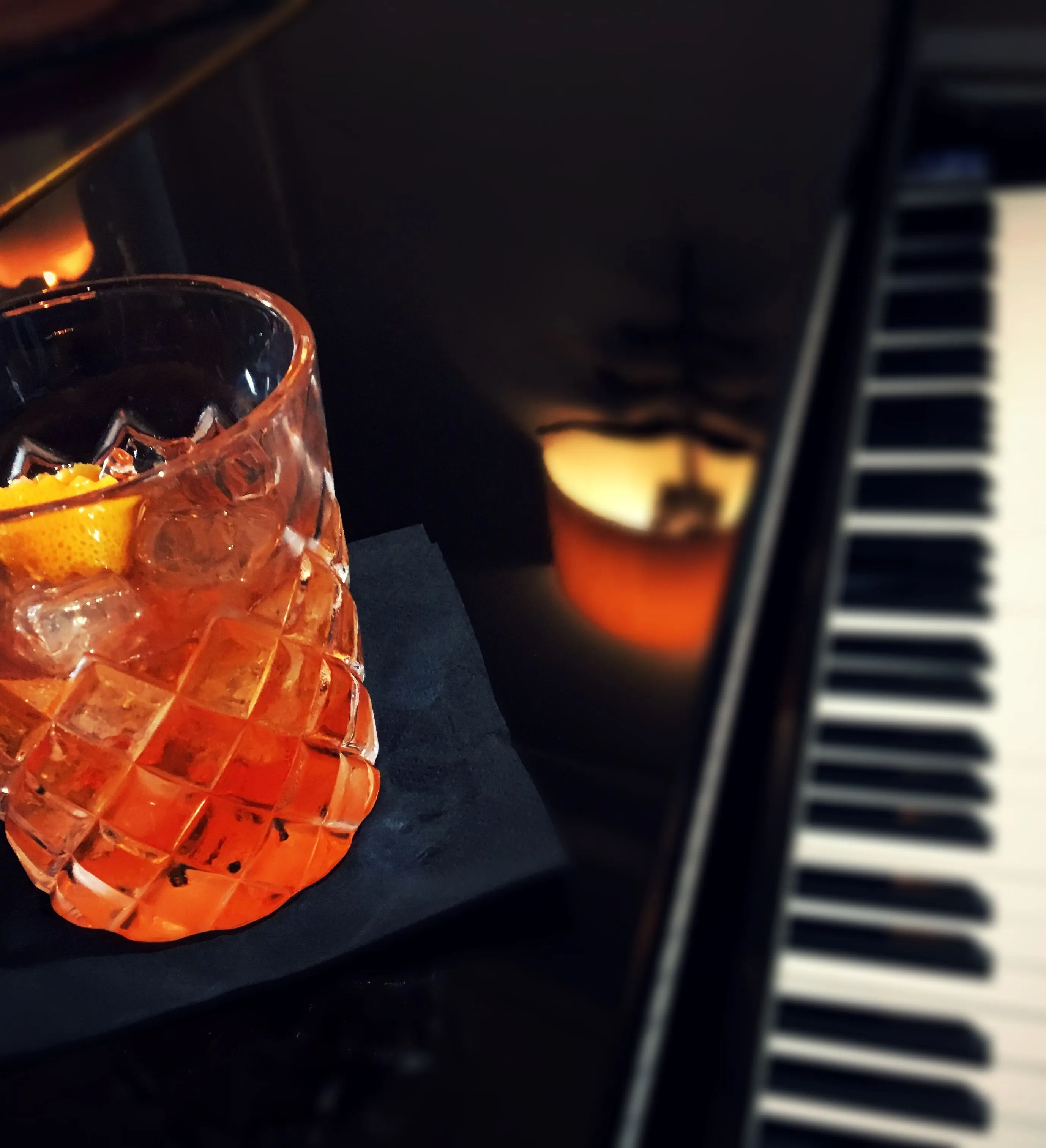 A glass of cocktail with an orange peel garnish on a black napkin, beside a piano keyboard.