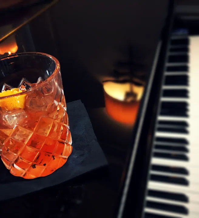 A glass of cocktail with an orange peel garnish on a black napkin, beside a piano keyboard.