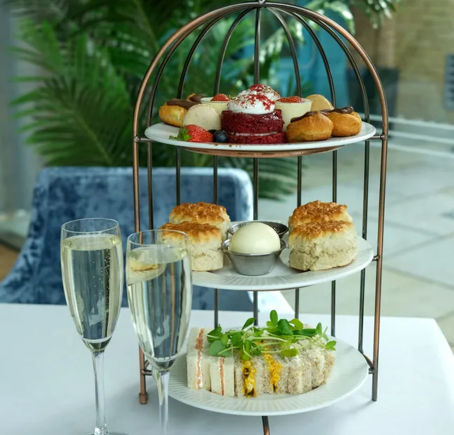 Three-tiered stand with assorted afternoon tea treats and two glasses of champagne on a white tablecloth.