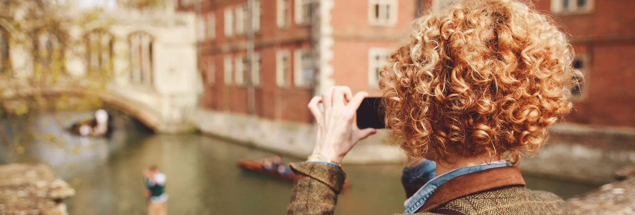 Person with curly hair photographs punts on the River Cam in Cambridge, standing on a stone bridge with historic college buildings in the background.