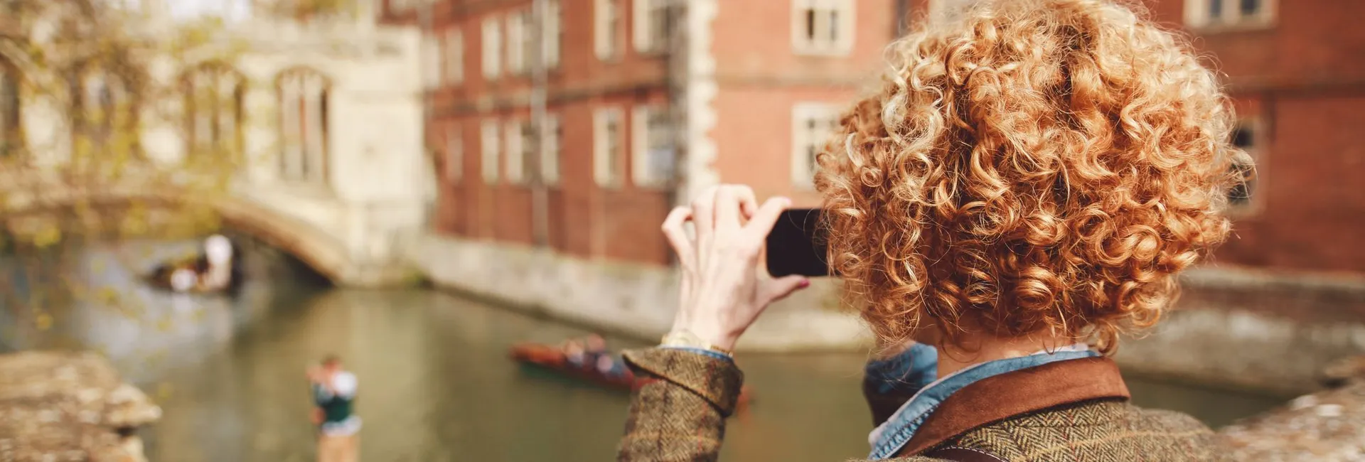 Person with curly hair photographs punts on the River Cam in Cambridge, standing on a stone bridge with historic college buildings in the background.
