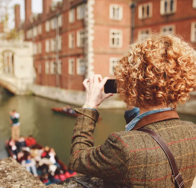 Person with curly hair photographs punts on the River Cam in Cambridge, standing on a stone bridge with historic college buildings in the background.
