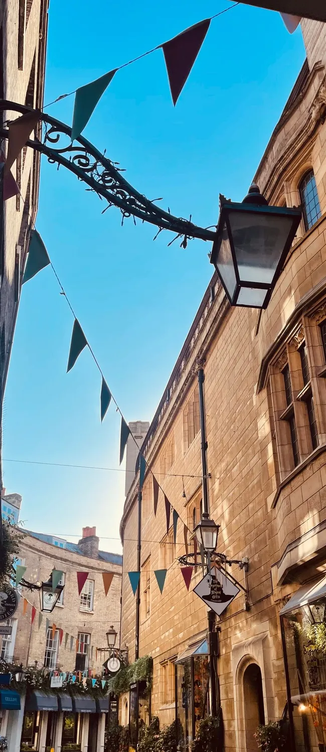 Narrow Cambridge street lined with historic stone buildings, festive bunting overhead, and shopfronts below under a clear blue sky.