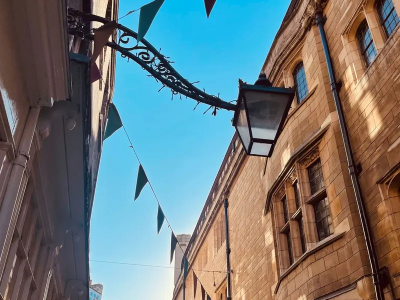 Narrow Cambridge street lined with historic stone buildings, festive bunting overhead, and shopfronts below under a clear blue sky.
