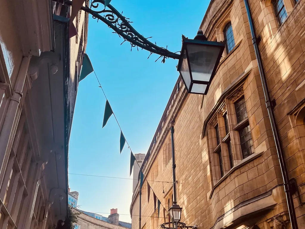 Narrow Cambridge street lined with historic stone buildings, festive bunting overhead, and shopfronts below under a clear blue sky.