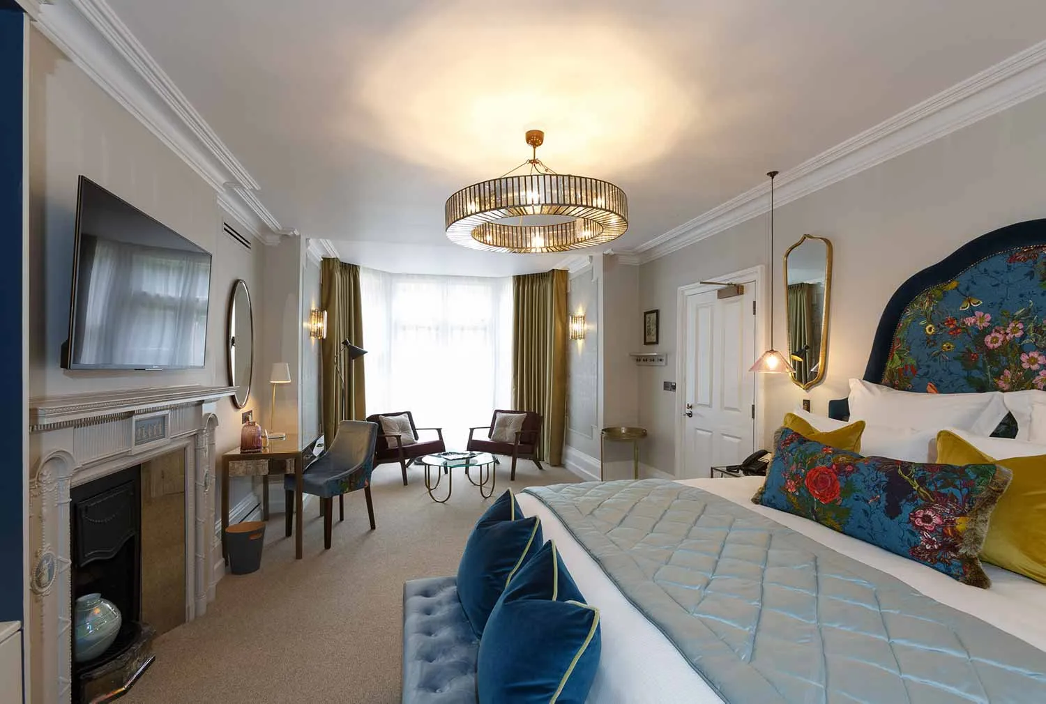 The Delphinium feature room at Gonville Hotel, with a floral headboard, velvet accents, chandelier lighting, seating area, desk, and fireplace.
