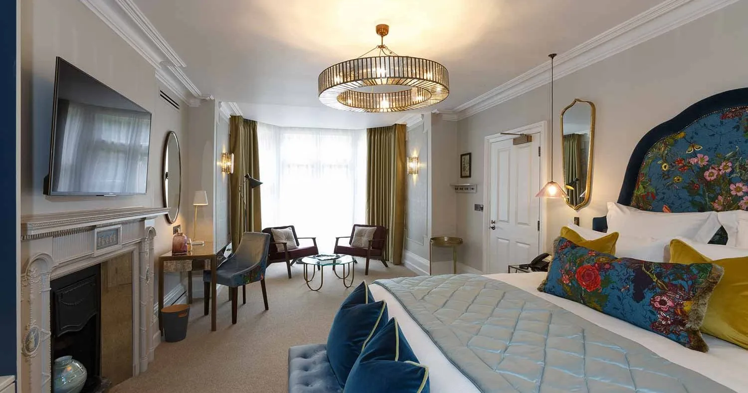 The Delphinium feature room at Gonville Hotel, with a floral headboard, velvet accents, chandelier lighting, seating area, desk, and fireplace.