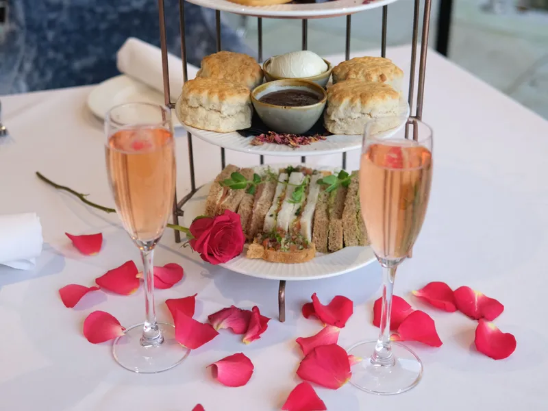 Three-tiered afternoon tea stand with cakes, scones, sandwiches, and two glasses of sparkling rosé on a table decorated with pink rose petals.