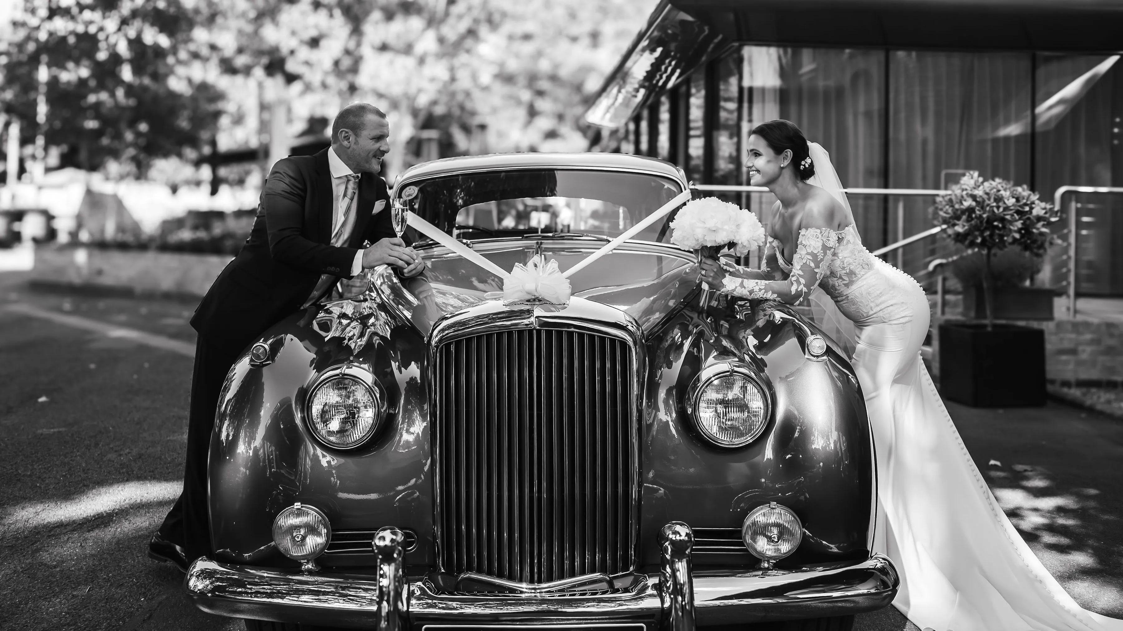 Bride and groom smiling at each other over the hood of a classic car decorated for a wedding.