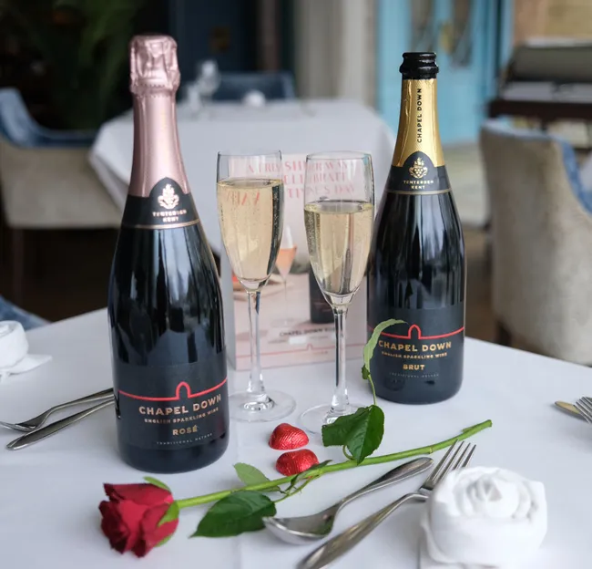 Chapel Down sparkling wine bottles with two filled glasses, a red rose, and heart-shaped chocolates on a restaurant table