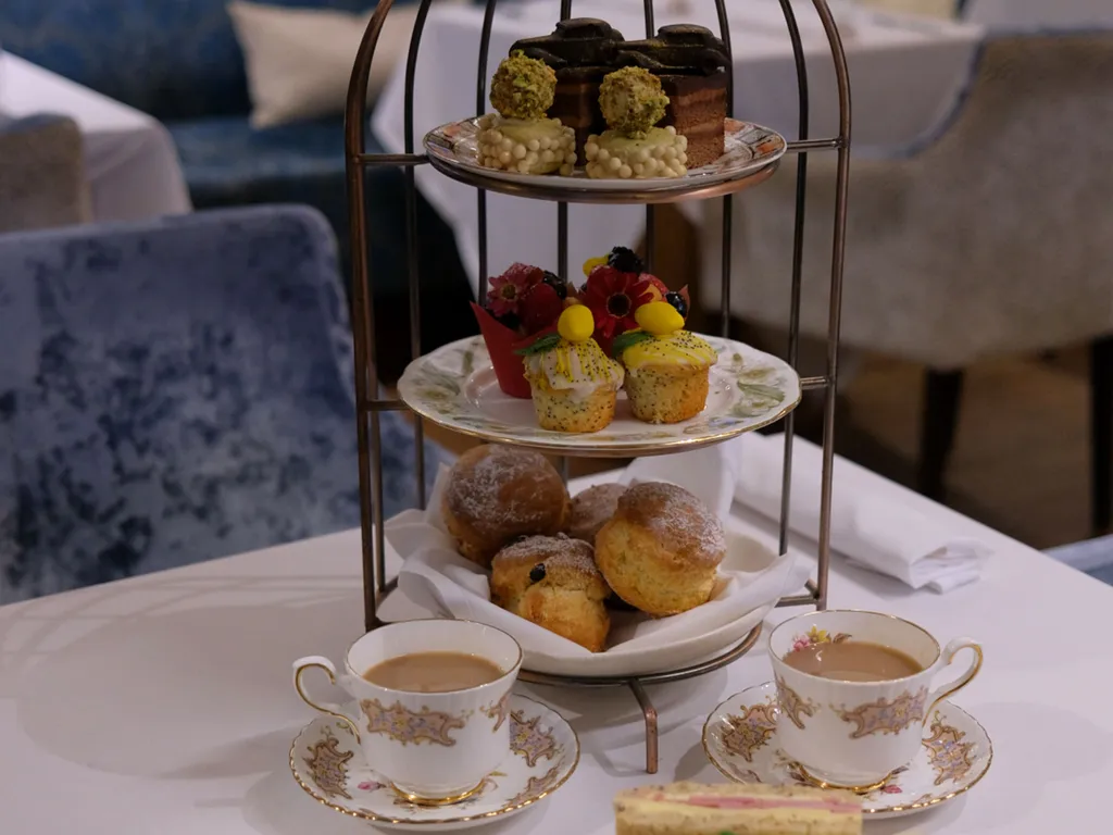 A traditional afternoon tea setup with a three-tiered stand holding pastries, scones, and desserts, surrounded by cups of tea and a plate of assorted finger sandwiches.