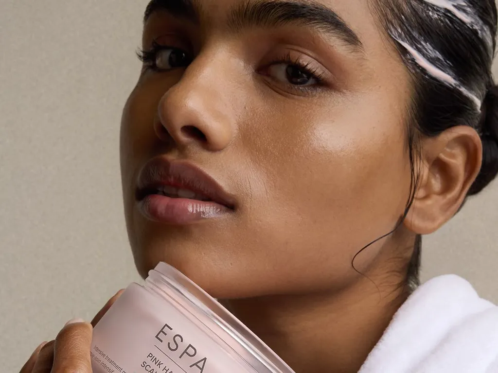 Close-up of a woman in a white robe holding an open ESPA Pink Hair & Scalp Mud jar, with product visible in her hair.