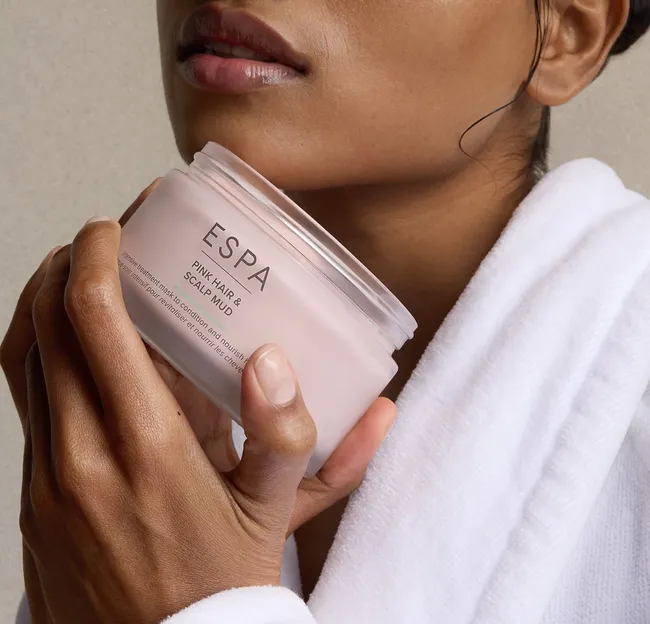 Close-up of a woman in a white robe holding an open ESPA Pink Hair & Scalp Mud jar, with product visible in her hair.