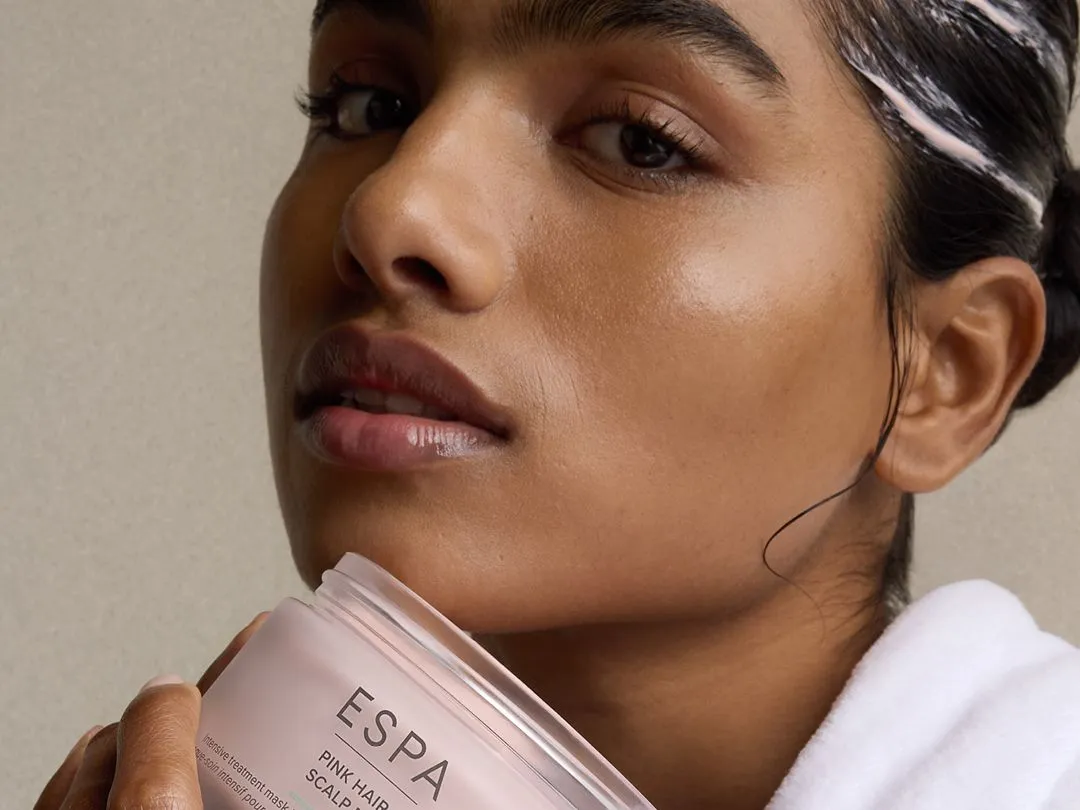 Close-up of a woman in a white robe holding an open ESPA Pink Hair & Scalp Mud jar, with product visible in her hair.