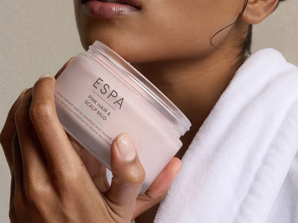 Close-up of a woman in a white robe holding an open ESPA Pink Hair & Scalp Mud jar, with product visible in her hair.