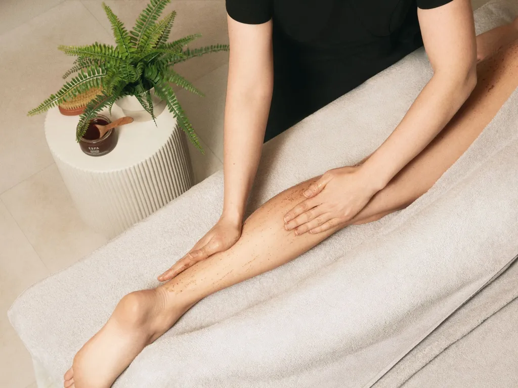 Therapist exfoliating a client’s leg with body scrub on a massage table