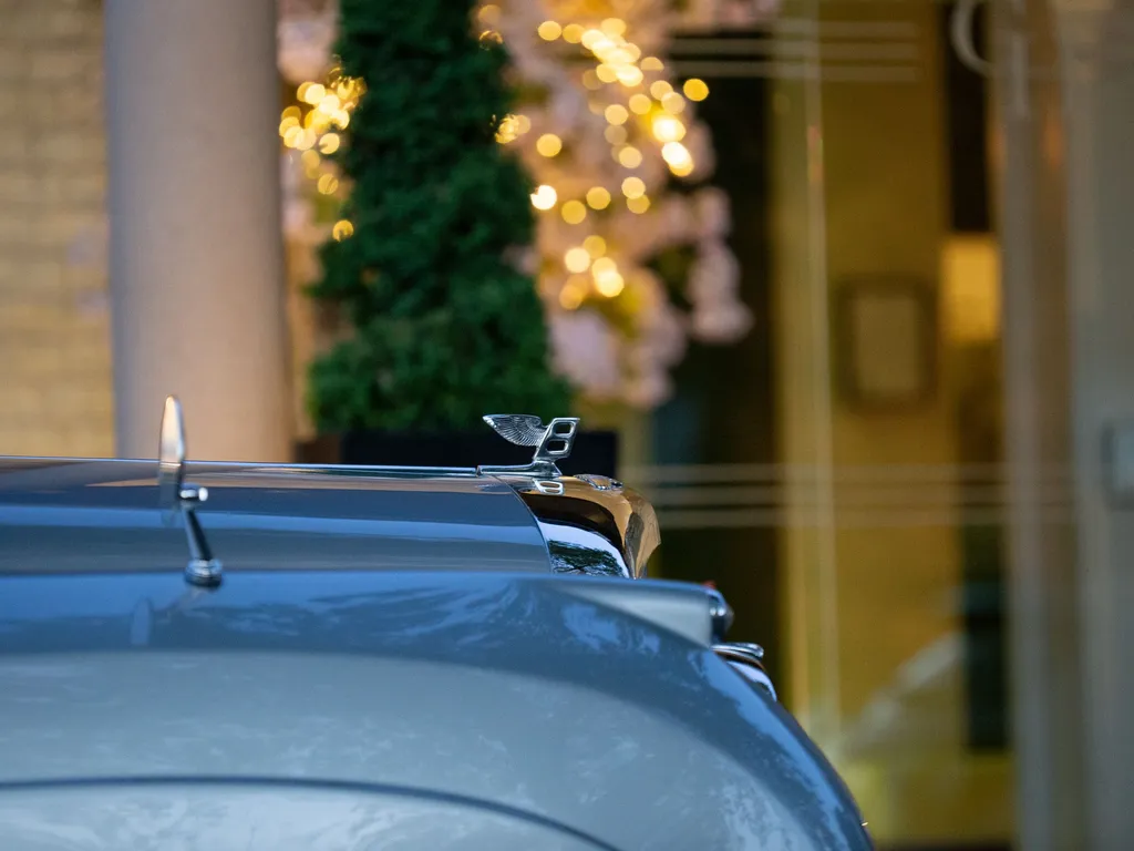 Close-up of a Bentley bonnet mascot on a grey car with warm festive lights blurred in the background