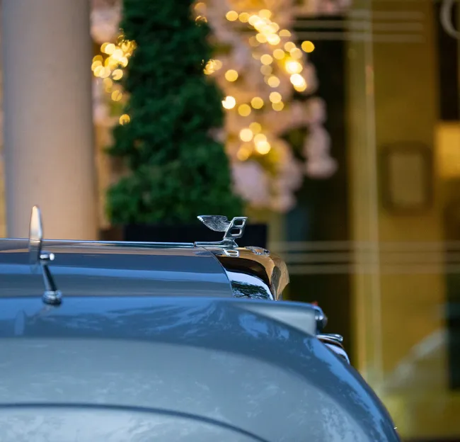 Close-up of a Bentley bonnet mascot on a grey car with warm festive lights blurred in the background