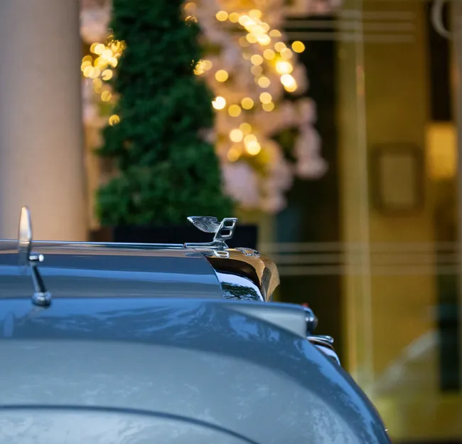 Close-up of a Bentley bonnet mascot on a grey car with warm festive lights blurred in the background