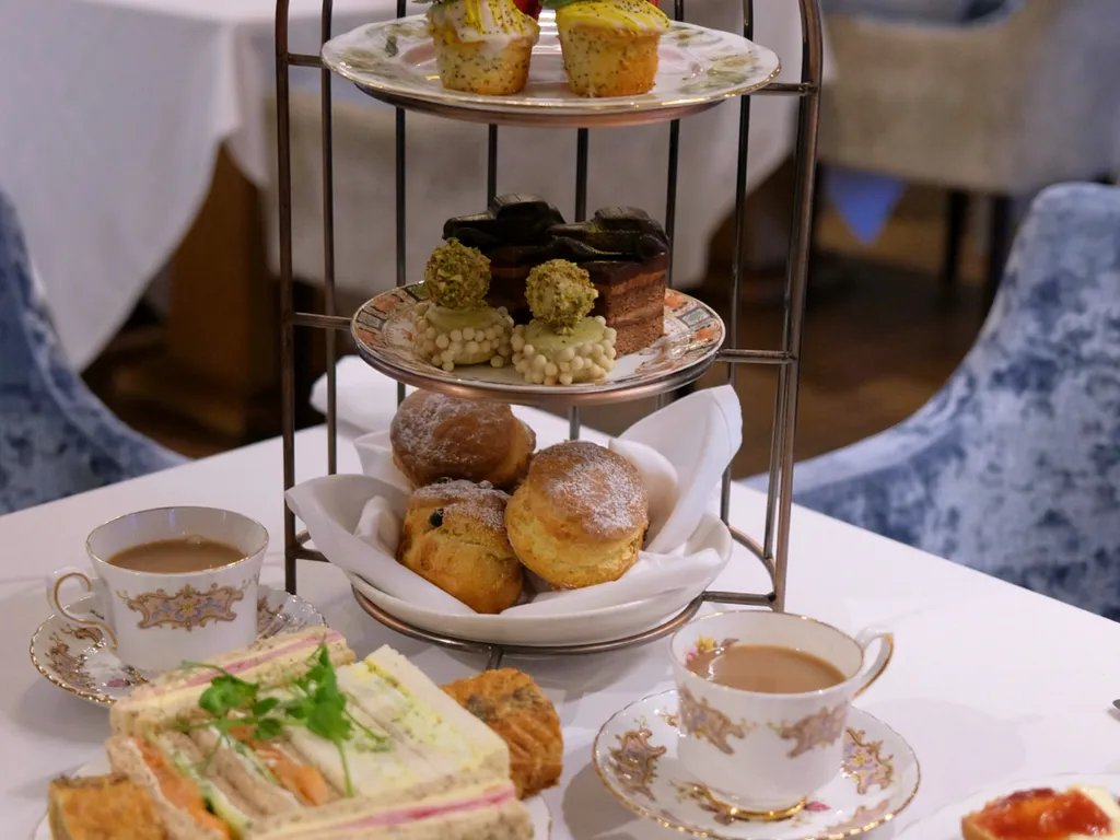 Three-tier afternoon tea stand with cakes, scones, sandwiches and teacups on a white table