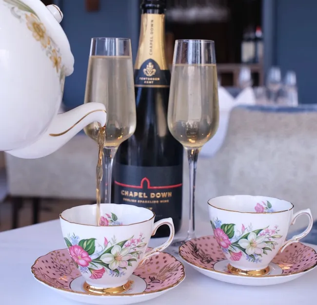 Champagne being poured from a floral teapot into a teacup beside two flutes and a bottle on a table in a restaurant