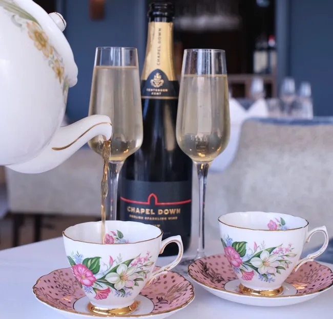 Champagne being poured from a floral teapot into a teacup beside two flutes and a bottle on a table in a restaurant