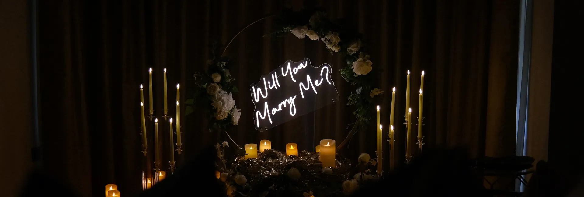 Romantic candlelit proposal set-up with illuminated 'Will You Marry Me?' sign and floral arch