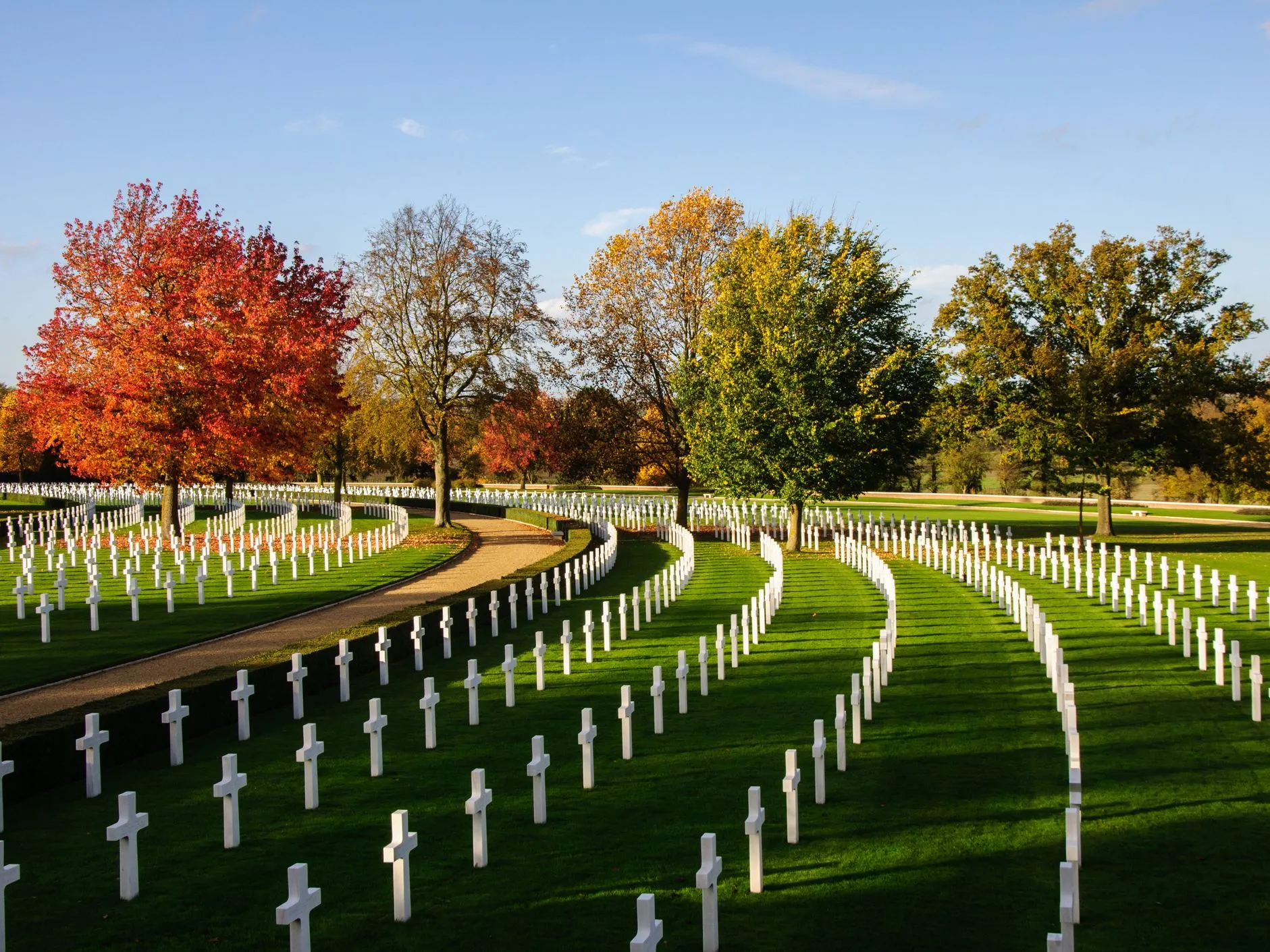 Rows of white crosses curve across the American Cemetery in Cambridge, framed by autumn trees and a clear blue sky.