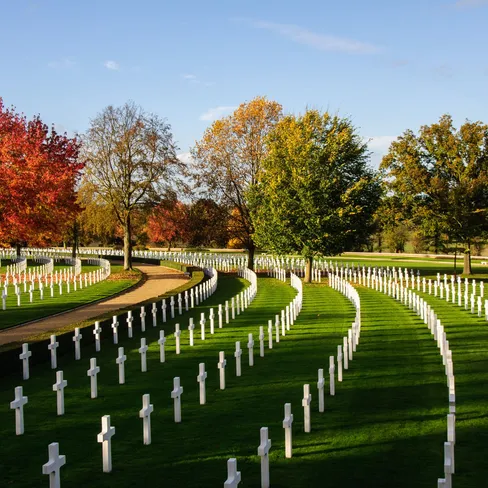Rows of white crosses curve across the American Cemetery in Cambridge, framed by autumn trees and a clear blue sky.