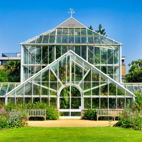 Glasshouse at the University of Cambridge Botanic Garden, framed by flower beds, benches and bright green lawn under a clear blue sky.
